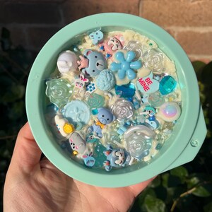 Könnte beinhalten: Ein mintgrüner Behälter gefüllt mit einer Vielzahl von blauen und weißen dekorativen Elementen. Der Inhalt umfasst Teddybären, Blumen und herzförmige Akzente, mit den Worten "BE MINE" auf einem Stück. Der Behälter wird in einer Hand gehalten.