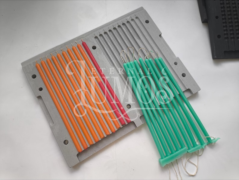 May include: A close-up shot of a candle-making mold with several orange, red, and green candles. The candles are long and thin, and they are arranged in rows within the mold. The mold is gray and has a rectangular shape. The text "ETERNAL MOS" is visible.