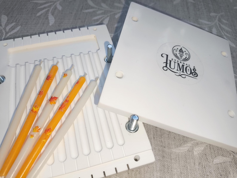 May include: White candle making mold with several white and orange candles. The orange candles have a leaf design. The mold has the text "LUMOS Eternal &" on the lid.