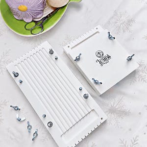 May include: Two white wooden weaving looms with metal hardware and wing nuts. One loom has vertical grooves, the other has the Lumos logo. A purple flower-shaped candle holder, green plate, scissors, and a spool of thread are in the background.