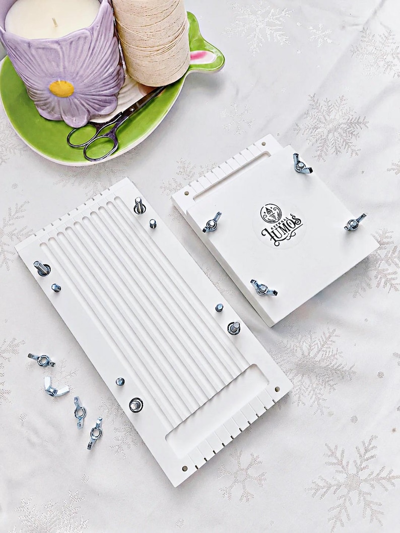 May include: Two white wooden weaving looms with metal hardware and wing nuts. One loom has vertical grooves, the other has the Lumos logo. A purple flower-shaped candle holder, green plate, scissors, and a spool of thread are in the background.