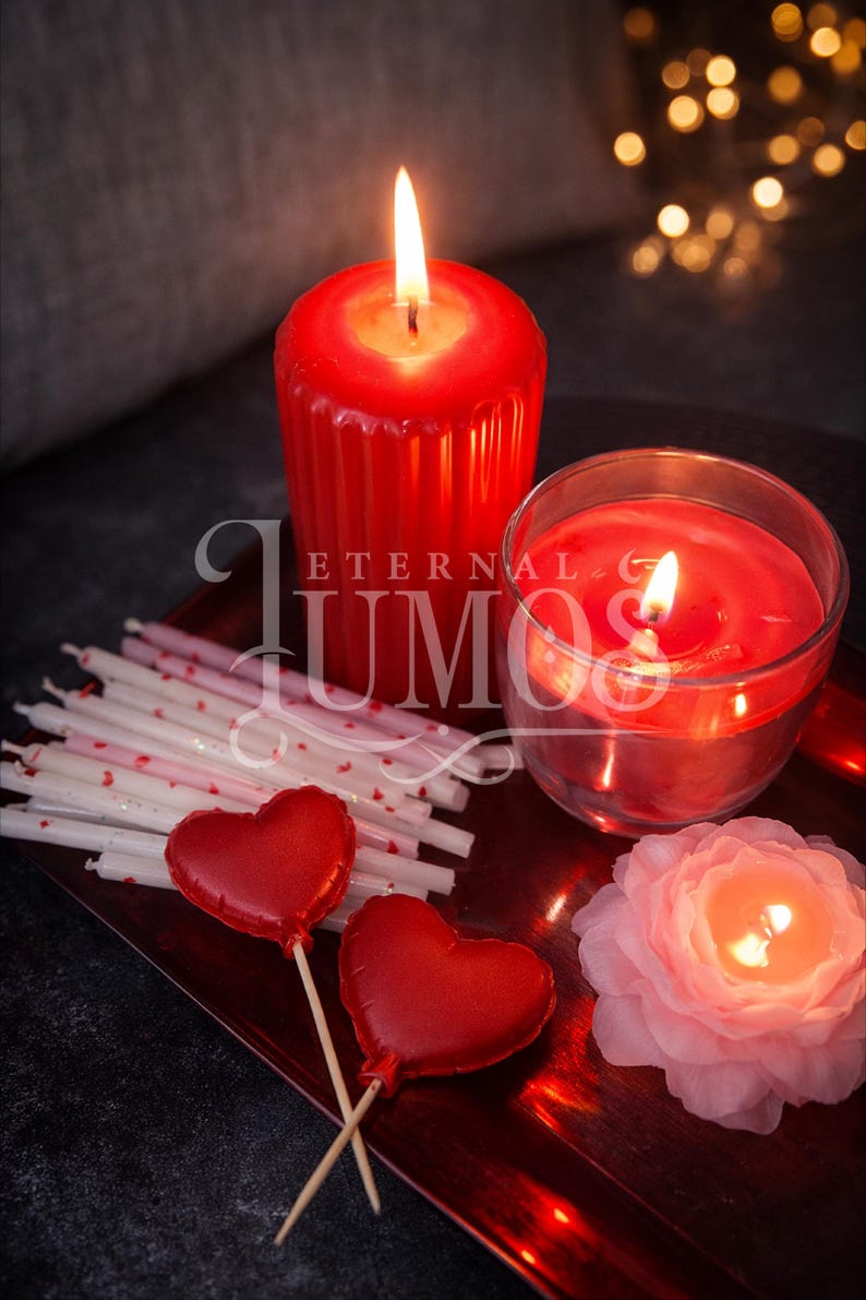 May include: A romantic still life featuring lit red candles of various shapes, including a large pillar candle and a glass-enclosed candle. Heart-shaped lollipops and a flower-shaped candle are also present, all arranged on a red tray.