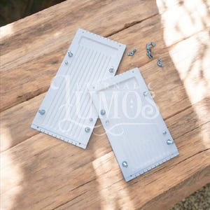 May include: Two white rectangular objects with parallel grooves and metal hardware are displayed on a wooden surface. Several loose screws and bolts are scattered nearby. The objects appear to be components of a larger item.
