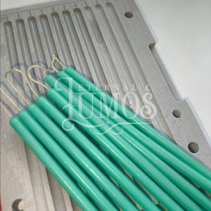 May include: A collection of teal-colored taper candles with wicks, resting on a gray candle mold. The candles are cylindrical and have a smooth finish. The mold has multiple slots for candle creation. The image includes the text "ETERNAL LUMOS".