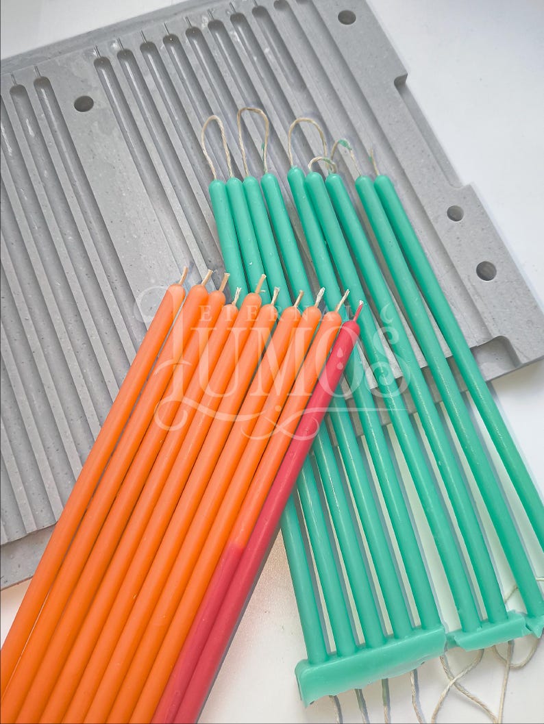 May include: A collection of colorful, slender candles in shades of orange, pink, and teal, arranged near a gray candle mold. The candles are of varying lengths and have wicks. The teal candles are grouped together, while the orange and pink candles are in a separate bundle.