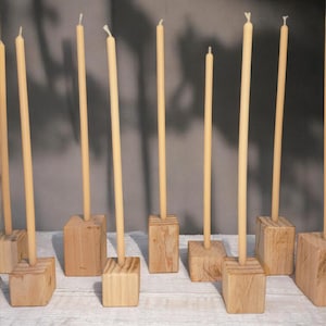 Peut inclure: Plusieurs bougies fines de couleur crème, chacune étant placée dans un petit socle carré en bois. Les bougies sont disposées sur une surface blanche bordée de dentelle, créant une présentation simple et élégante. Les tons de bois naturel et la couleur douce des bougies créent une esthétique chaleureuse.