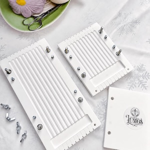 May include: White weaving loom set with metal hardware, including wing nuts and screws. The set includes two rectangular looms, a small square piece with a logo, and additional hardware. The looms have vertical grooves and are designed for weaving projects.