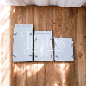 May include: Three white rectangular boxes of varying sizes, each with decorative silver hardware, are arranged on a wooden surface. The boxes are closed and appear to be empty. The word "LUMOS" is partially visible on the middle box.