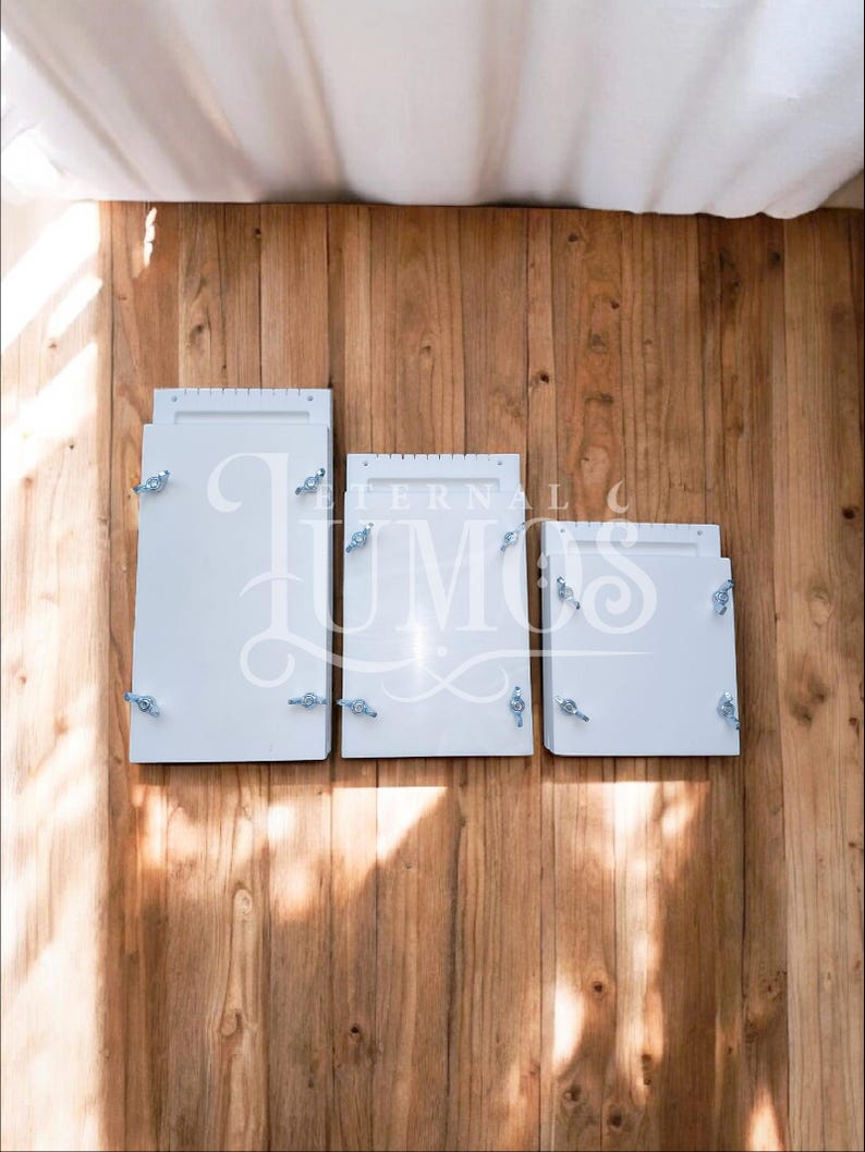 May include: Three white rectangular boxes of varying sizes, each with decorative silver hardware, are arranged on a wooden surface. The boxes are closed and appear to be empty. The word "LUMOS" is partially visible on the middle box.
