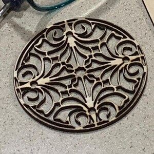 Könnte beinhalten: Ein runder Holz-Untersetzer mit einem dekorativen Laser-Schnittmuster. Das Design zeigt komplizierte Wirbel und Muster, die einen zarten und eleganten Look erzeugen.