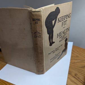 May include: An open vintage book titled "Keeping Fit - Health". The cover features a silhouette of a person exercising. The book's spine displays the title and author's name. The book is on a wooden surface.