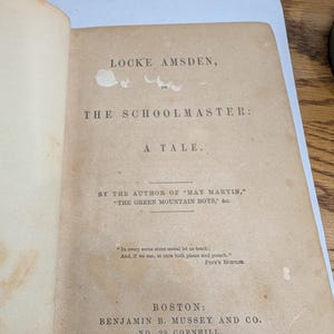 May include: Open book page with text: "LOCKE AMSDEN, ON THE SCHOOLMASTER: A TALE." The author is listed as the author of "MAY MARTIN" and "THE GREEN MOUNTAIN BOYS." Published in Boston in 1853.