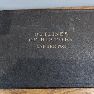 May include: A vintage, black book titled "OUTLINES OF HISTORY LABBERTON" in gold lettering. The book has a worn, textured cover and is resting on a wooden surface. The book's binding shows signs of age.