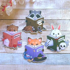 Könnte beinhalten: Vier gestanzte Aufkleber mit einem braunen Hamster, der ein Romance-Buch liest, einem grauen Waschbären mit einem Mystery Who?-Buch, einem orangefarbenen Fuchs mit einem Fantasy-Buch und einem weißen Kaninchen mit einem Horror-Buch samt Totenkopf.
