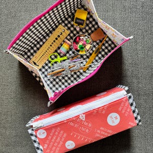 May include: Two zippered pouches, one with a black and white checked fabric exterior and a pink zipper, and the other with a red fabric exterior with white text and a white zipper. The pouch with the checked fabric is open and contains various sewing and knitting supplies, including a ruler, scissors, yarn, and a needle.