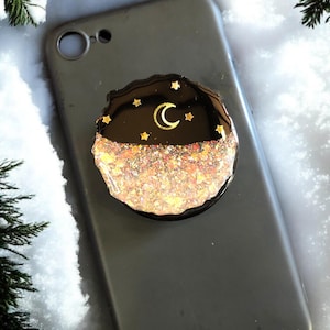 May include: A grey mobile phone with a decorative phone grip attached. The grip has a black top with gold stars and a crescent moon, and a glitter-filled bottom.