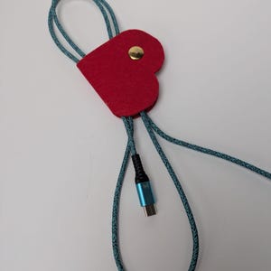 Könnte beinhalten: Ein roter herzförmiger Kabelorganizer mit einem goldfarbenen Druckknopfverschluss. Der Organizer ist an einem teal-schwarzen geflochtenen Kabel mit einem blauen USB-C-Anschluss befestigt. Das Kabel ist geschlungen.
