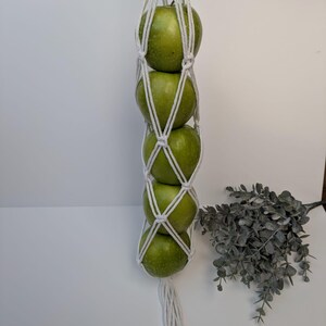 Op de afbeelding: Een witte macramé fruitmand met vier groene appels. Het geweven ontwerp omhult de appels en creëert een decoratieve en functionele opbergoplossing. De houder is opgehangen en toont de appels tegen een witte achtergrond.