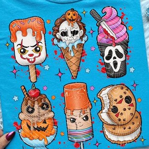 May include: A light blue t-shirt featuring cartoon ice cream cones with horror movie themes. The ice cream cones have various designs, including a clown, a skull, and a cookie sandwich. The design is surrounded by stars and blood splatters.