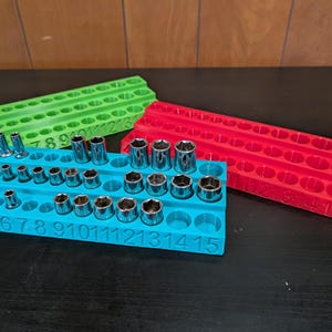 May include: Three colorful socket organizers in green, red, and blue. The blue organizer holds various sockets, with numbers printed on the front. The green and red organizers are empty. The background is a wooden wall and a black surface.