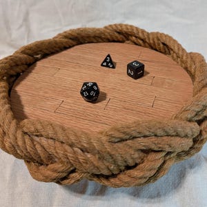 May include: A wooden dice tray with a rope border. The tray has a light brown wood finish and is designed to hold dice. There are three black dice on the tray, showing the numbers 1, 2, and 3.