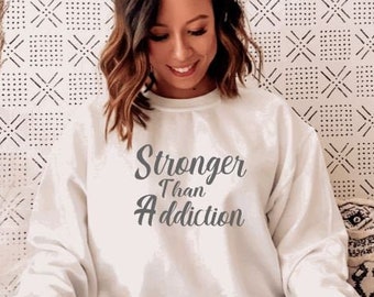 以下が含まれることがあります： グレーの文字で「Stronger Than Addiction」と書かれた白いスウェットシャツ。モデルはスウェットシャツを着てソファに座っています。
