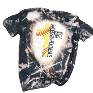 May include: A black t-shirt with a bleach-splattered design. A yellow cross with a softball graphic is centered on the shirt. The text "All I need is a little bit of softball and a whole lot of Jesus" is printed on the cross.