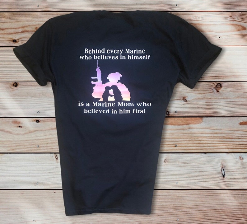 Peut inclure: T-shirt noir avec un imprim&eacute; graphique blanc repr&eacute;sentant une silhouette de Marine et de maman Marine. Le texte dit "Derri&egrave;re chaque Marine qui croit en lui-m&ecirc;me se trouve une maman Marine qui a cru en lui en premier."