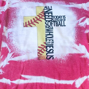May include: A red and white tie-dye t-shirt with a yellow baseball cross design. The text on the shirt reads "All I Need is a little bit of Softball and a Whole Lot of Jesus".
