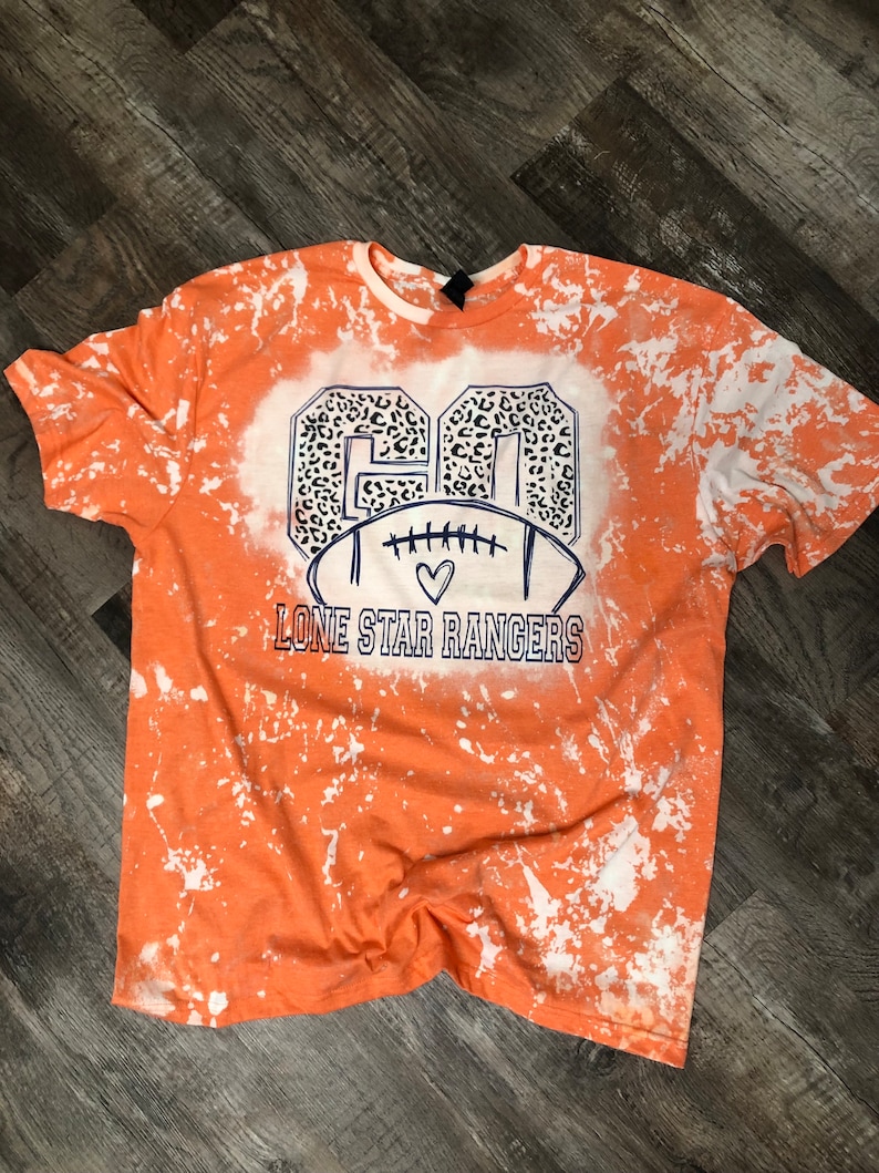 K&ouml;nnte beinhalten: Orangefarbenes, gebleichtes T-Shirt mit einem wei&szlig;en und schwarzen Leopardenmuster-Fu&szlig;ball-Design. Der Text "Lone Star Rangers" ist unter dem Fu&szlig;ball gedruckt.