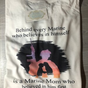 Peut inclure: T-shirt blanc avec un motif graphique noir et blanc. Le graphique repr&eacute;sente une silhouette de Marine tenant un fusil avec un coucher de soleil en arri&egrave;re-plan. Le texte "Behind every Marine who believes in himself is a Marine Mom who believed in him first" est imprim&eacute; sur le t-shirt.