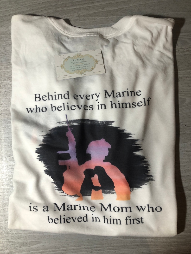 Peut inclure: T-shirt blanc avec un motif graphique noir et blanc. Le graphique repr&eacute;sente une silhouette de Marine tenant un fusil avec un coucher de soleil en arri&egrave;re-plan. Le texte "Behind every Marine who believes in himself is a Marine Mom who believed in him first" est imprim&eacute; sur le t-shirt.