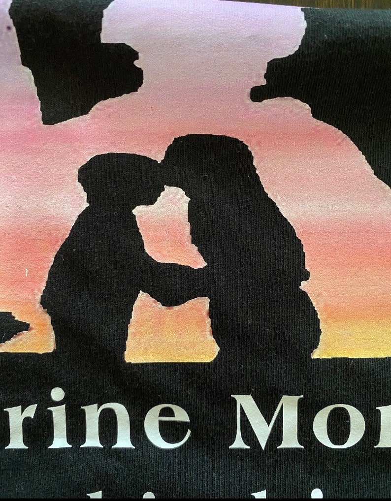 Peut inclure: T-shirt noir avec une silhouette de deux personnes qui s'embrassent devant un coucher de soleil rose, orange et jaune. Le texte "Marine Mom" est imprim&eacute; sous la silhouette.