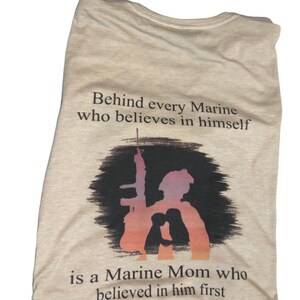 Peut inclure: T-shirt beige avec une silhouette d'un Marine tenant un fusil et une m&egrave;re de Marine derri&egrave;re lui. Le texte dit "Behind every Marine who believes in himself is a Marine Mom who believed in him first."