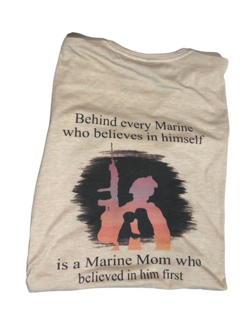 Peut inclure: T-shirt beige avec une silhouette d'un Marine tenant un fusil et une m&egrave;re de Marine derri&egrave;re lui. Le texte dit "Behind every Marine who believes in himself is a Marine Mom who believed in him first."