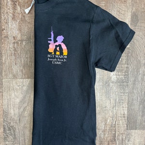 Peut inclure: T-shirt noir avec une silhouette d'un soldat tenant un fusil et un enfant. Le texte "SGT. MAJOR Joseph Sosa Jr. USMC" est imprim&eacute; sur le t-shirt.