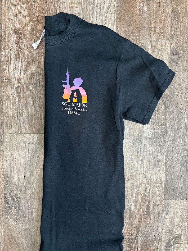 Peut inclure: T-shirt noir avec une silhouette d'un soldat tenant un fusil et un enfant. Le texte "SGT. MAJOR Joseph Sosa Jr. USMC" est imprim&eacute; sur le t-shirt.