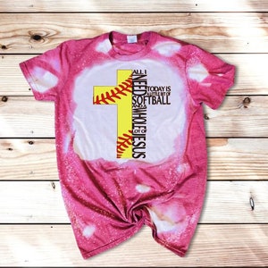 May include: Pink bleached t-shirt with a yellow softball cross design. The text reads "All I need is a little bit of softball and a whole lot of Jesus".