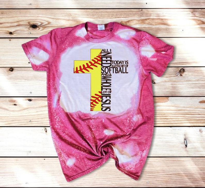 May include: Pink bleached t-shirt with a yellow softball cross design. The text reads "All I need is a little bit of softball and a whole lot of Jesus".