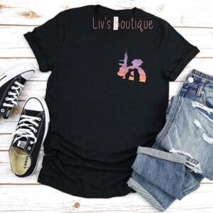 Peut inclure: T-shirt noir avec une silhouette de deux personnes debout devant un horizon urbain avec un fond d&eacute;grad&eacute; de coucher de soleil. Le texte "Liv's Boutique" est imprim&eacute; sur le t-shirt.