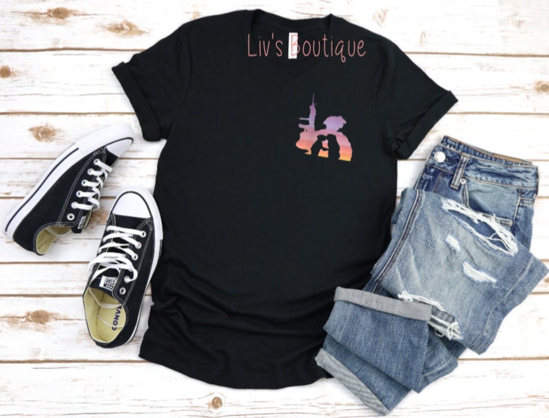 Peut inclure: T-shirt noir avec une silhouette de deux personnes debout devant un horizon urbain avec un fond d&eacute;grad&eacute; de coucher de soleil. Le texte "Liv's Boutique" est imprim&eacute; sur le t-shirt.