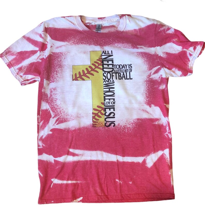 May include: Red and white tie-dye t-shirt with a yellow and red baseball cross design. The text on the shirt reads "All I need is a little bit of softball and a whole lot of Jesus".