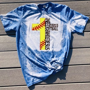 May include: A blue bleached t-shirt with a yellow softball cross design. The text on the shirt reads "All I need is a little bit of softball and a whole lot of Jesus."
