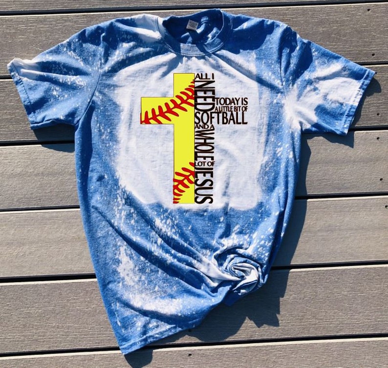 May include: A blue bleached t-shirt with a yellow softball cross design. The text on the shirt reads "All I need is a little bit of softball and a whole lot of Jesus."