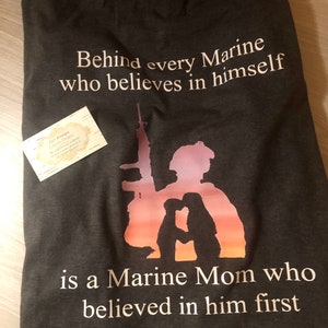 Peut inclure: Un t-shirt gris fonc&eacute; avec un motif graphique blanc. Le graphique repr&eacute;sente une silhouette d'un Marine et d'une m&egrave;re de Marine avec le texte "Behind every Marine who believes in himself is a Marine Mom who believed in him first."