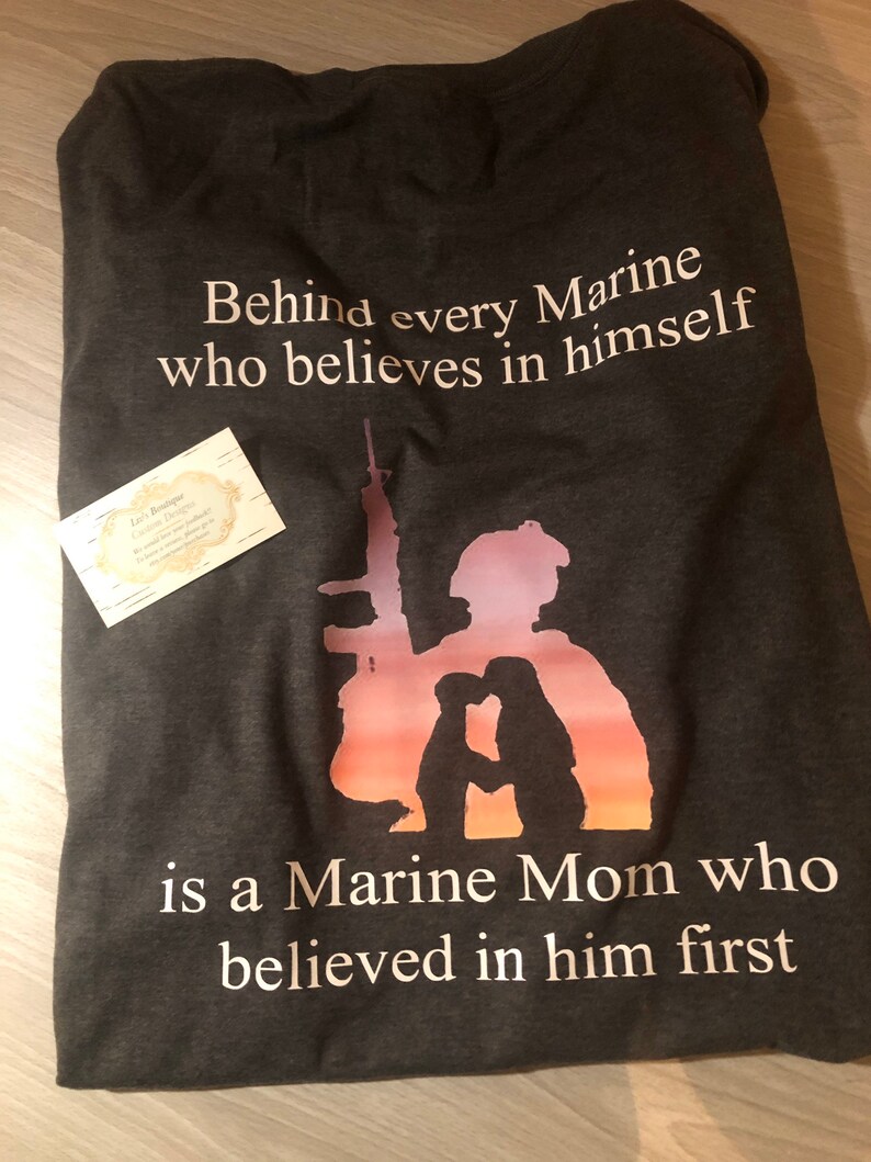 Peut inclure: Un t-shirt gris fonc&eacute; avec un motif graphique blanc. Le graphique repr&eacute;sente une silhouette d'un Marine et d'une m&egrave;re de Marine avec le texte "Behind every Marine who believes in himself is a Marine Mom who believed in him first."