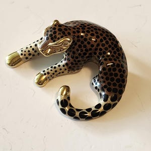 May include: A decorative leopard figurine in a resting pose. The figurine features a brown body with black spots, gold-colored paws, and a gold-colored head. The leopard's body is curved, and its tail is curled.