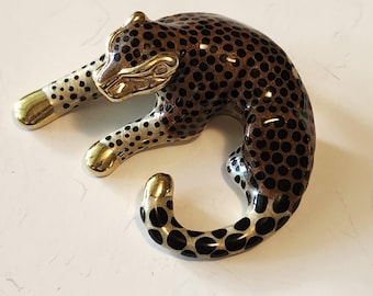 May include: A decorative leopard figurine in a resting pose. The figurine features a brown body with black spots, gold-colored paws, and a gold-colored head. The leopard's body is curved, and its tail is curled.