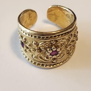 May include: A gold-colored ring with an open design. The ring features intricate floral and beaded patterns, with small purple gemstones set within the design. The ring is a wide band style.
