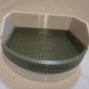 May include: A 3D-printed, semi-circular tray with a white base and a sage green rim and interior. The interior features a grid pattern. The tray has a raised back and side walls. The text "GE3D Designs" is visible on the rim.